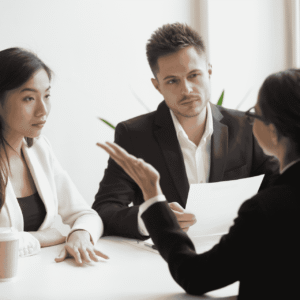 Three professionals sit at a white table in a meeting; a man in a suit gestures while two women listen to him over documents and coffee cups